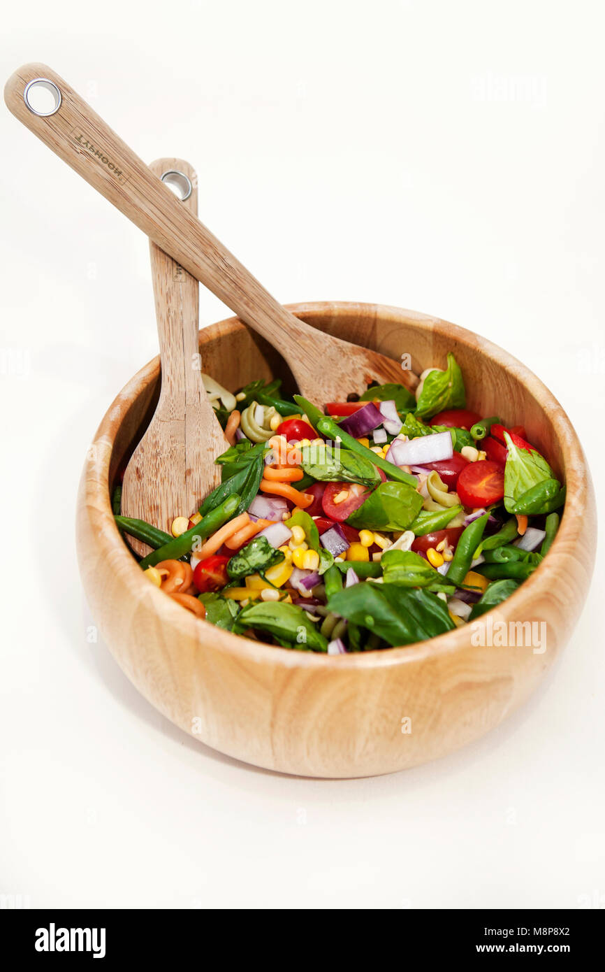 Pasta salad in a wooden bowl, with sweetcorn, onion, tomatoes and spinach. and with salad serving utensils. Stock Photo