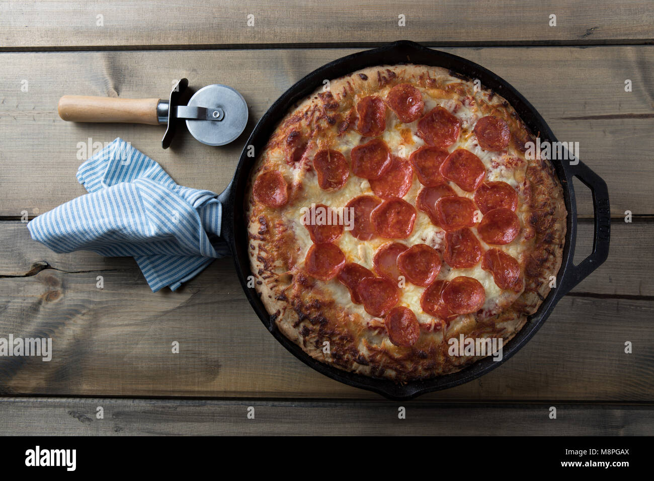 deep dish pizza in cast iron pan on rustic wooden table with pizza cutter and dish towel wrapped around handle Stock Photo
