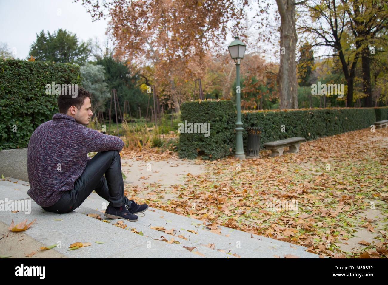 Thoughtful young man sitting on stairs in autumn park and looking away ...