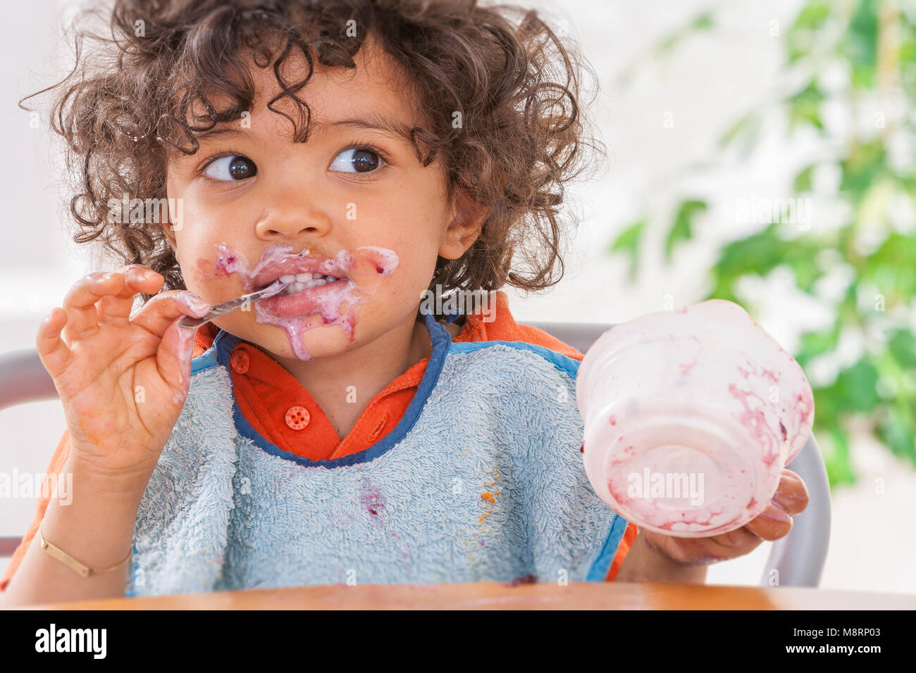 Toddler sitting in highchair and eating greek yogurt. Baby learning to eat and has yogurt on face and hair Stock Photo