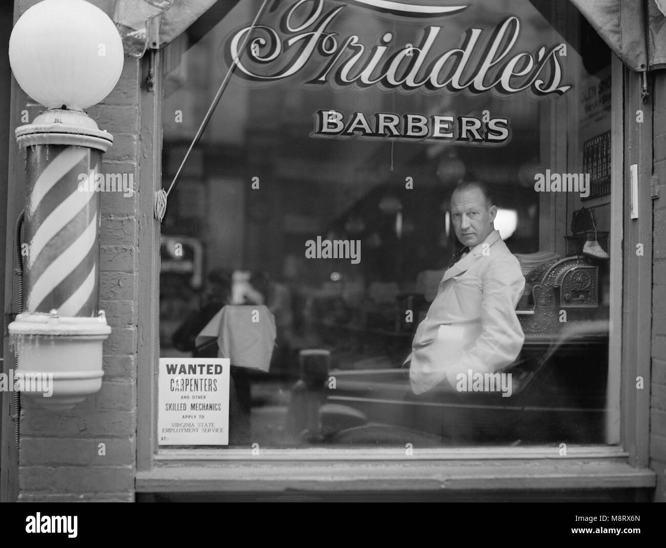 Portrait of Barber in Barber Shop Window, Help Wanted Sign Looking for Skilled Labor Resources Posted in Window by Virginia State Employment Service, Friddle's Barbers, Harrisonburg, Virginia, USA, John Vachon for Office for Emergency Management, May 1941 Stock Photo