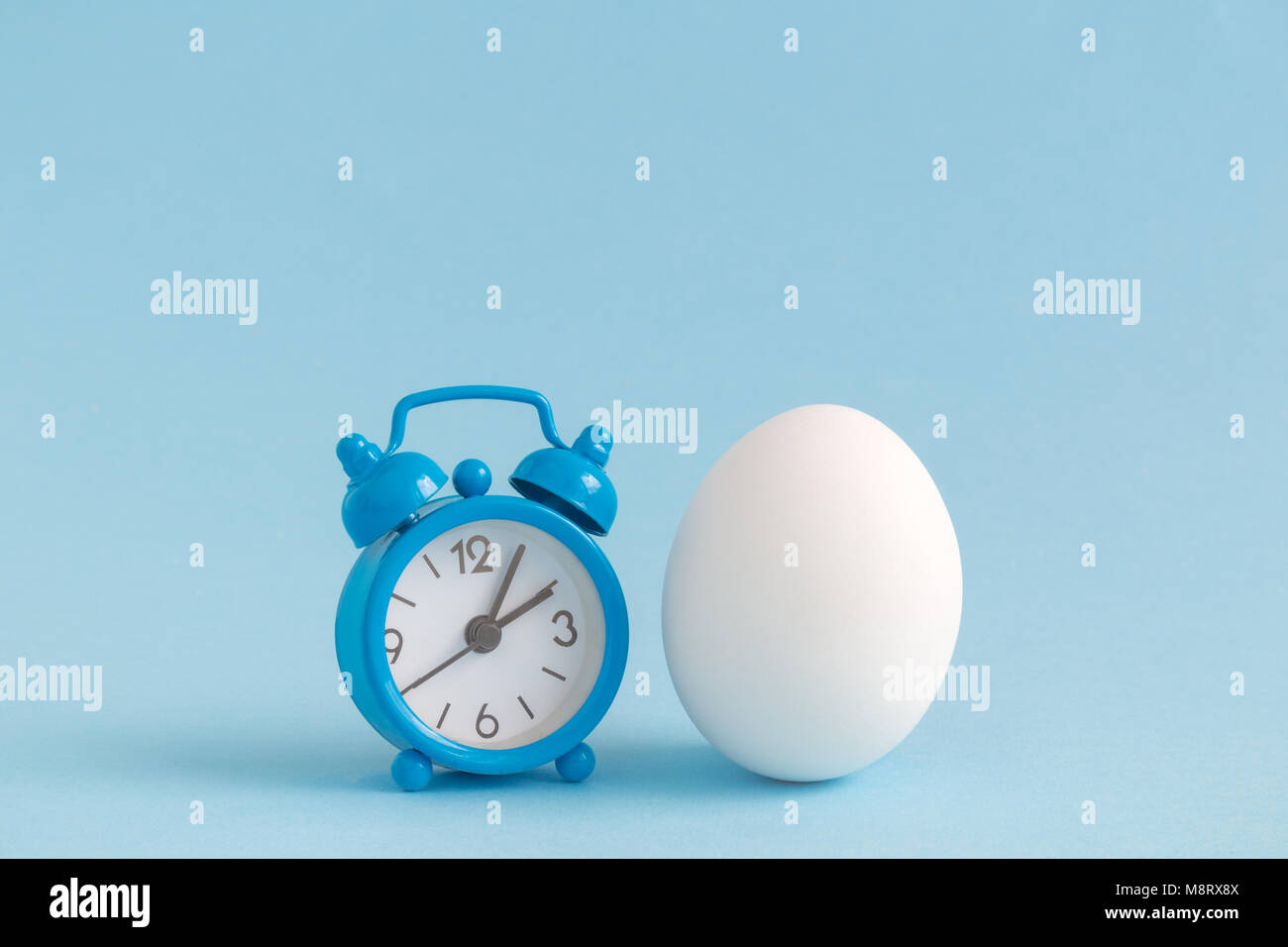 Small alarm clock and white egg on pastel blue background. Eastertime