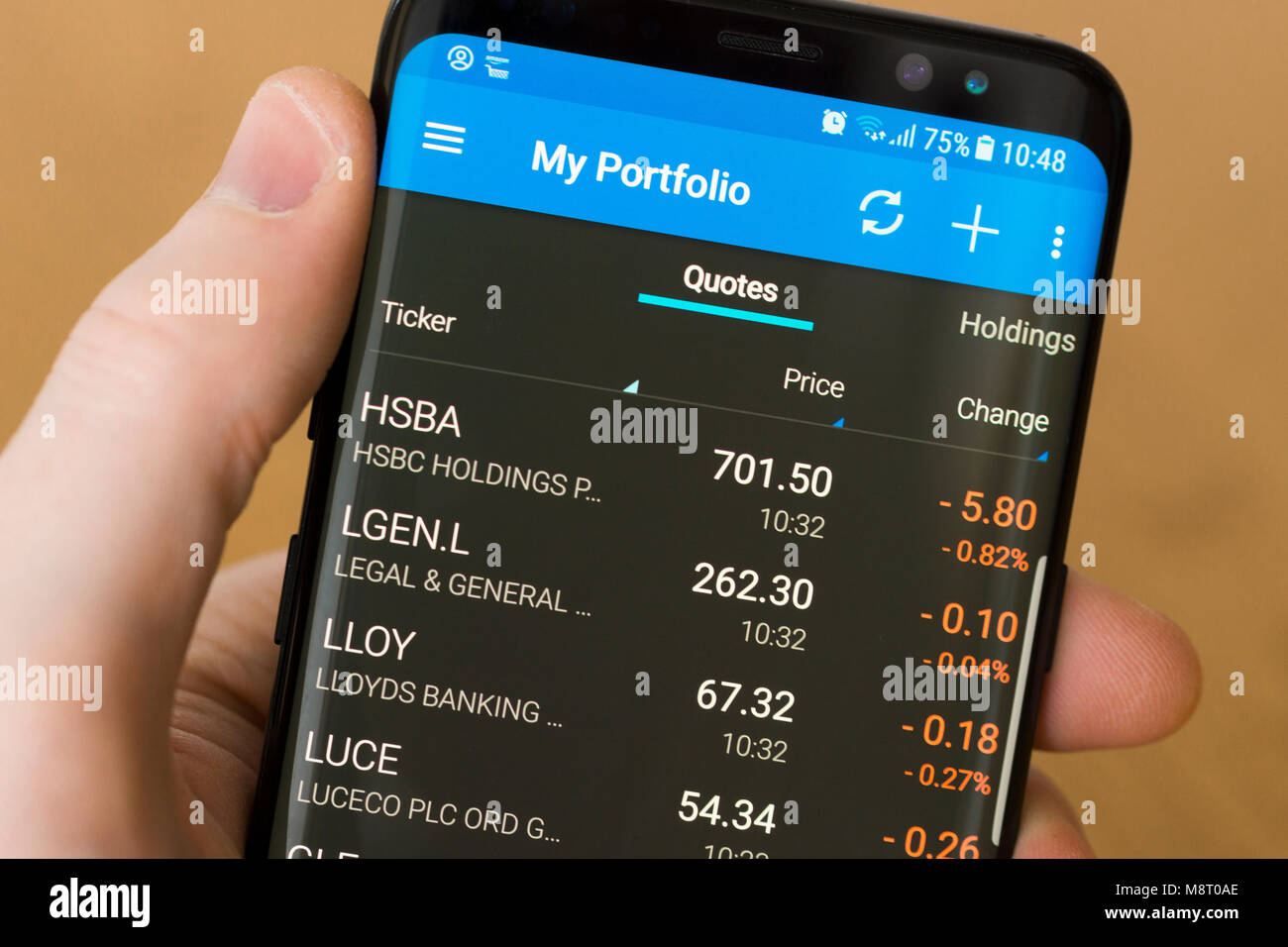 Someone checking their share prices on a smartphone. A man's hand holding a cellphone displaying stocks and shares all showing a loss Stock Photo