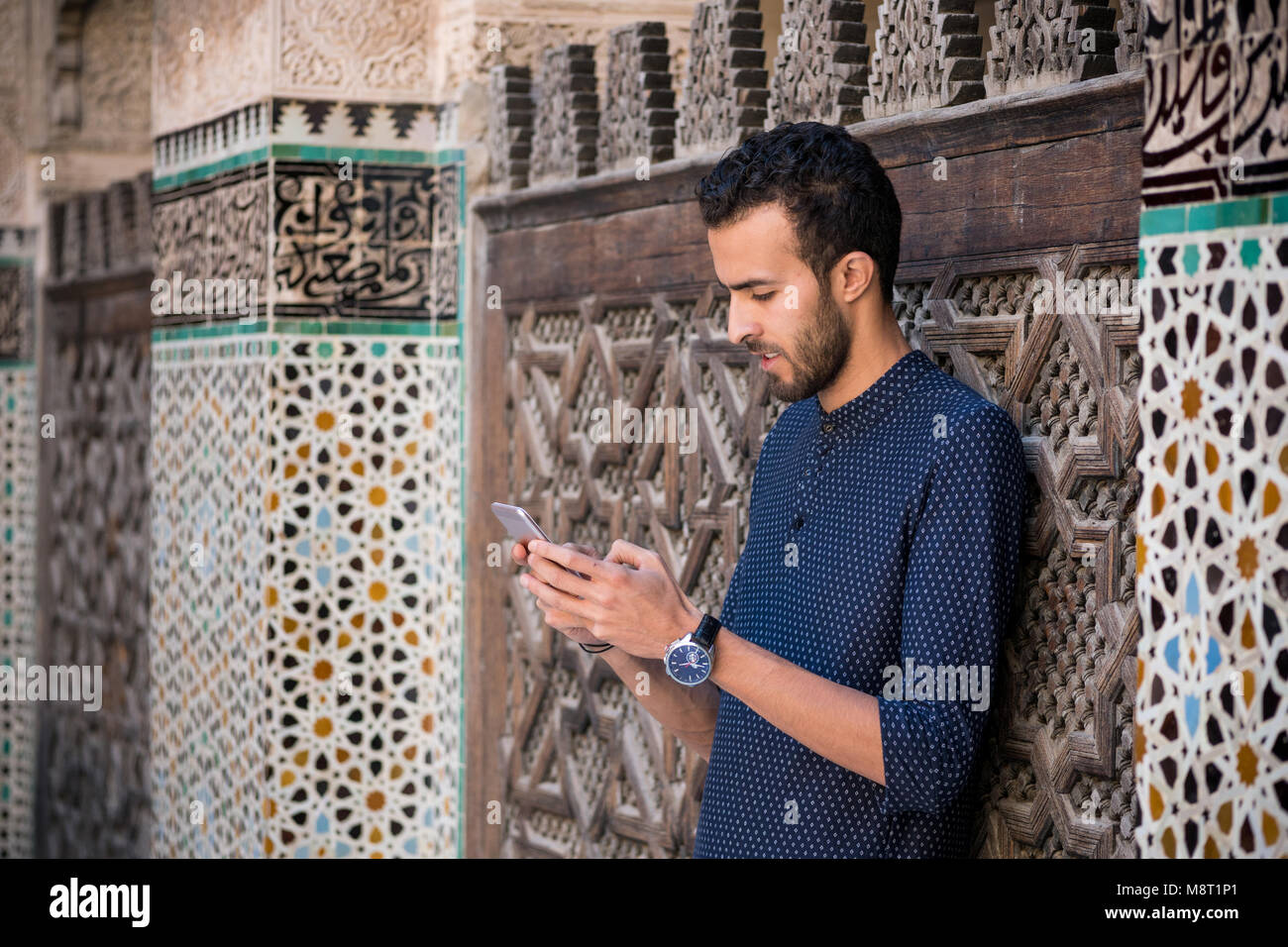 Young Muslim man using his mobile phone in casual clothing beside the wall with traditional Moroccan arabesque decoration Stock Photo