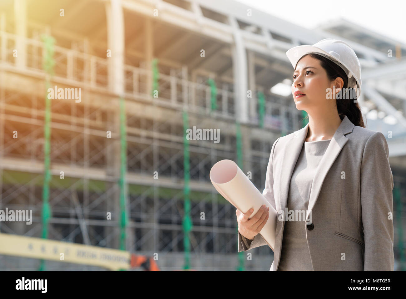 A female architect leader standing at the building site, staring forward with hope and confidence. Wearing a grey suit and holding the blueprint of th Stock Photo