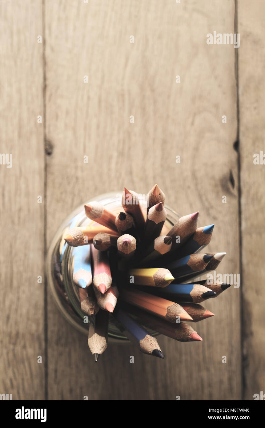 Overhead shot of multiple art pencils in many colours, grouped in a glass jar on oak wood planked table with copy space above. Filtered, retro style. Stock Photo