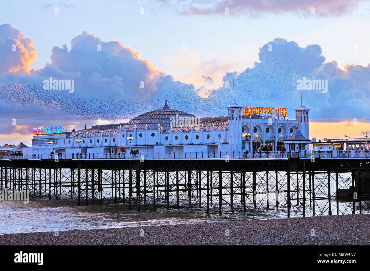 Brighton’s Palace Pier illuminated at early twilight, against a beautiful sunset with a murmuration of starlings swirling gracefully through the sky Stock Photo