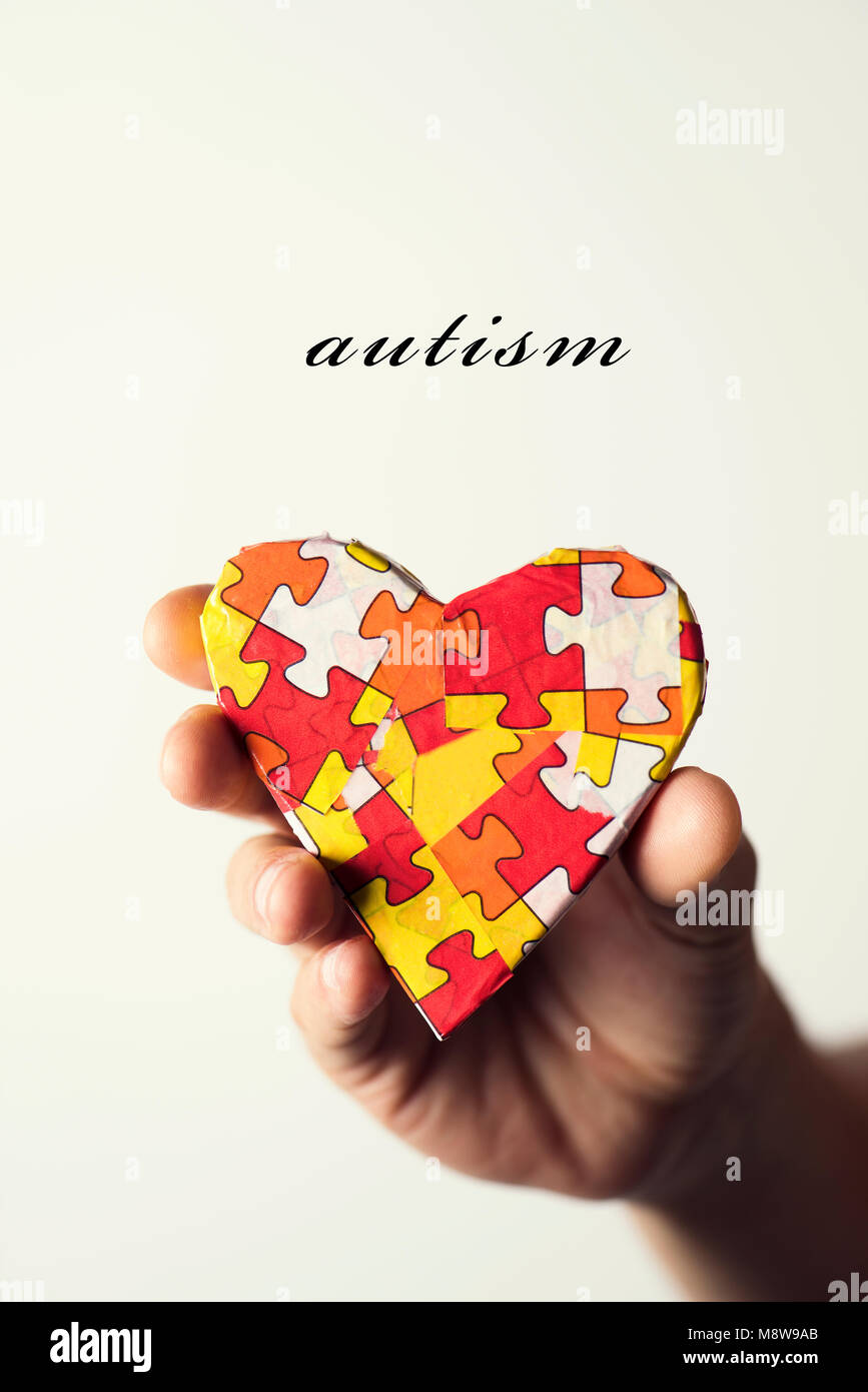 the word autism and a heart patterned with many puzzle pieces of different colors, symbol of the autism awareness, in the hand of a young caucasian ma Stock Photo