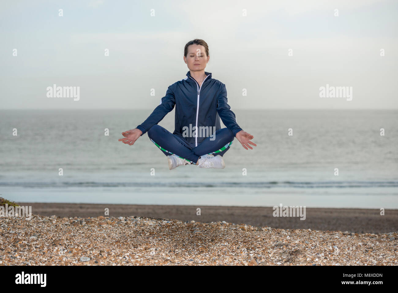 woman wearing a blue tracksuit in the lotus yoga position levitating above a beach Stock Photo