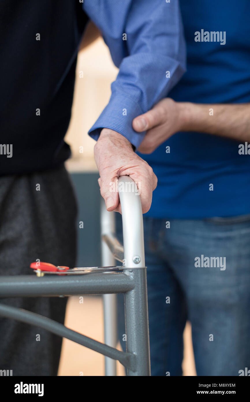 Senior Man's Hands On Walking Frame With Care Worker In Background Stock Photo