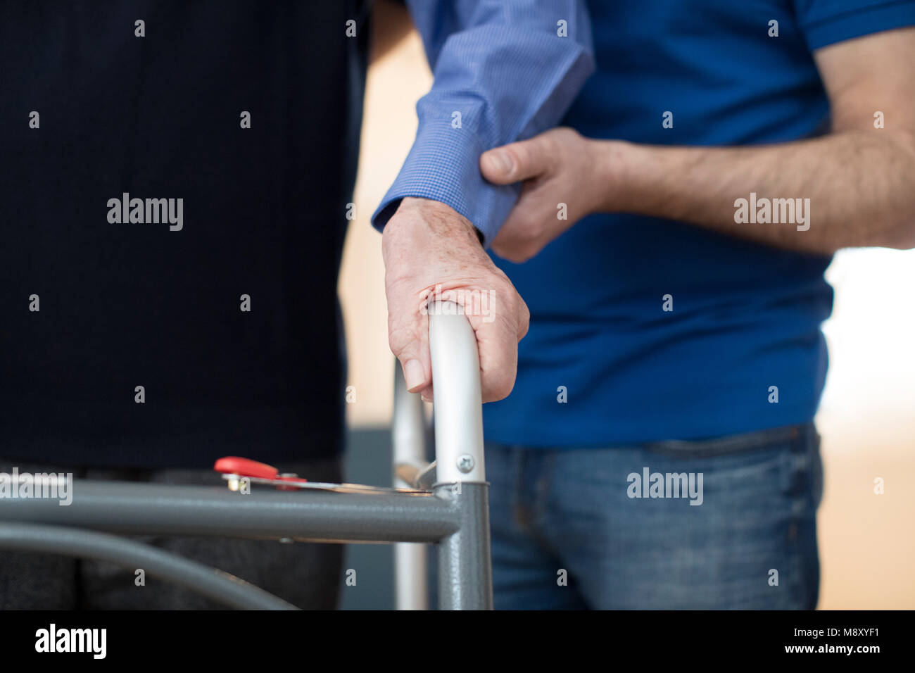 Senior Man's Hands On Walking Frame With Care Worker In Background Stock Photo