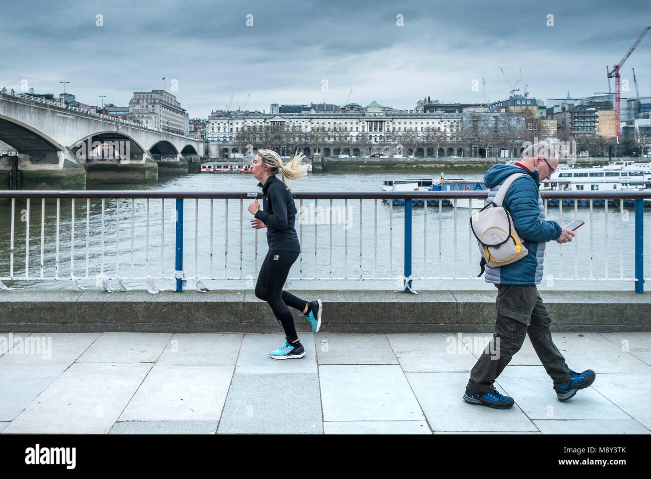 A woman running and a man walking on the South Bank in London. Stock Photo
