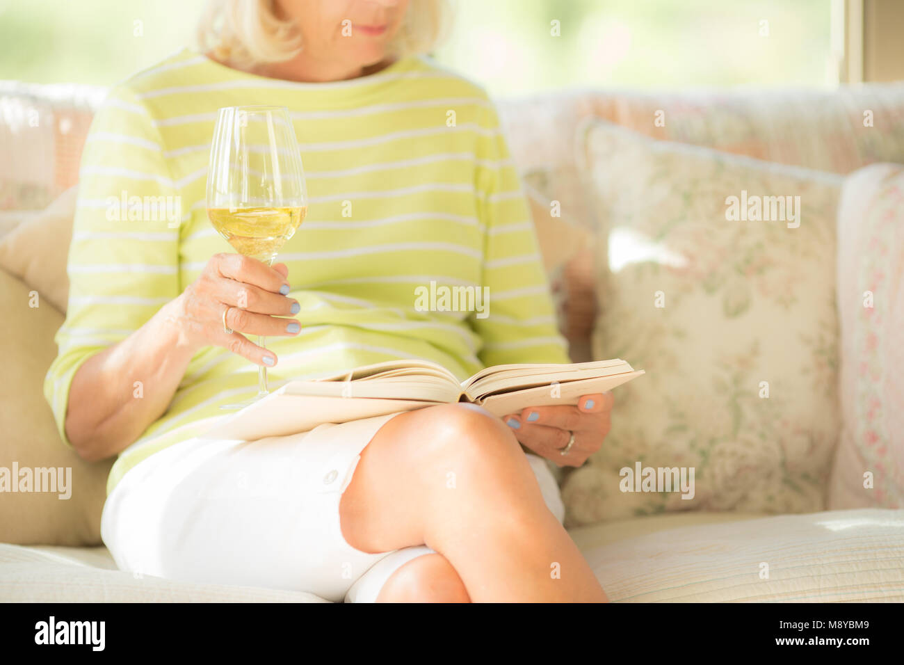 A mature woman drinking a glass of white wine while reading a book Stock Photo