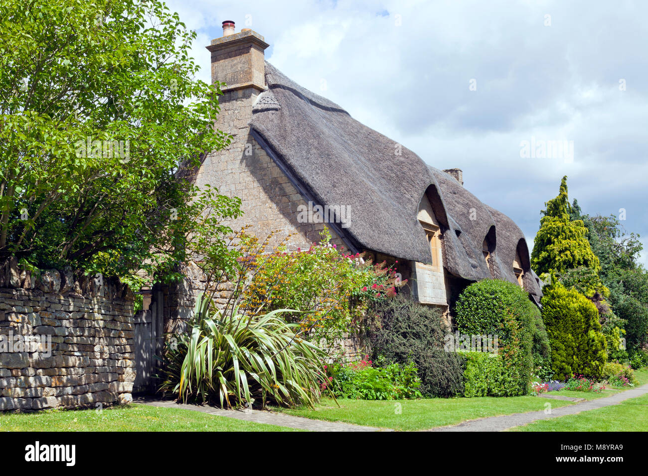 Village path along charming thatched roof English house in rural Cotswold countryside, with flowers, shrubs, trees front garden, , on a sunny summer d Stock Photo