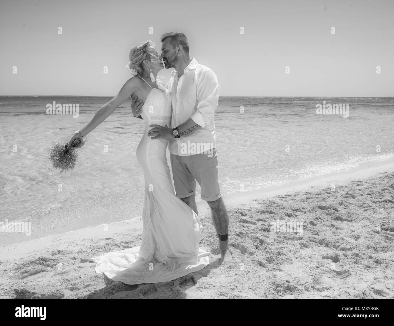 Beautiful married couple kissing at a tropical beach paradise on wedding day in gown dress with ocean view black and white photo Stock Photo