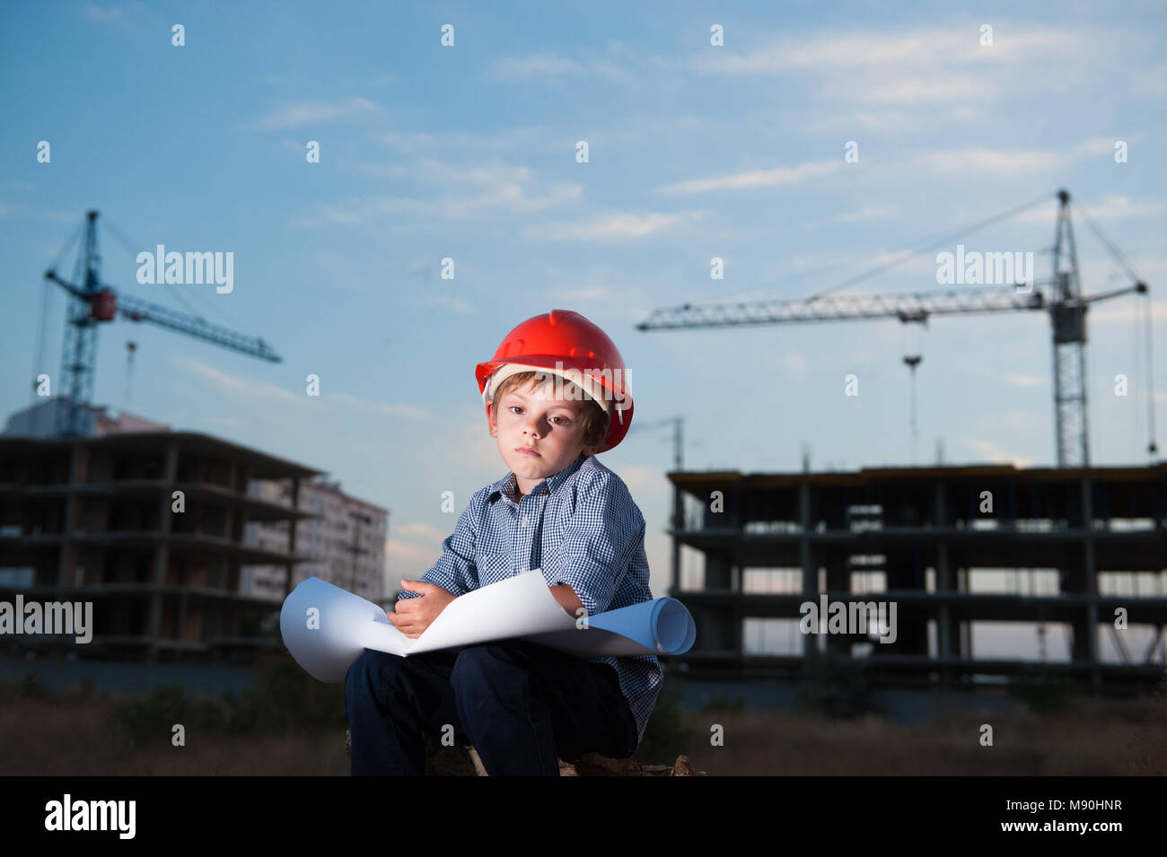 sad small child in casque sitting on construction site with paper plan in hands on background of crane and building in summer sunset Stock Photo