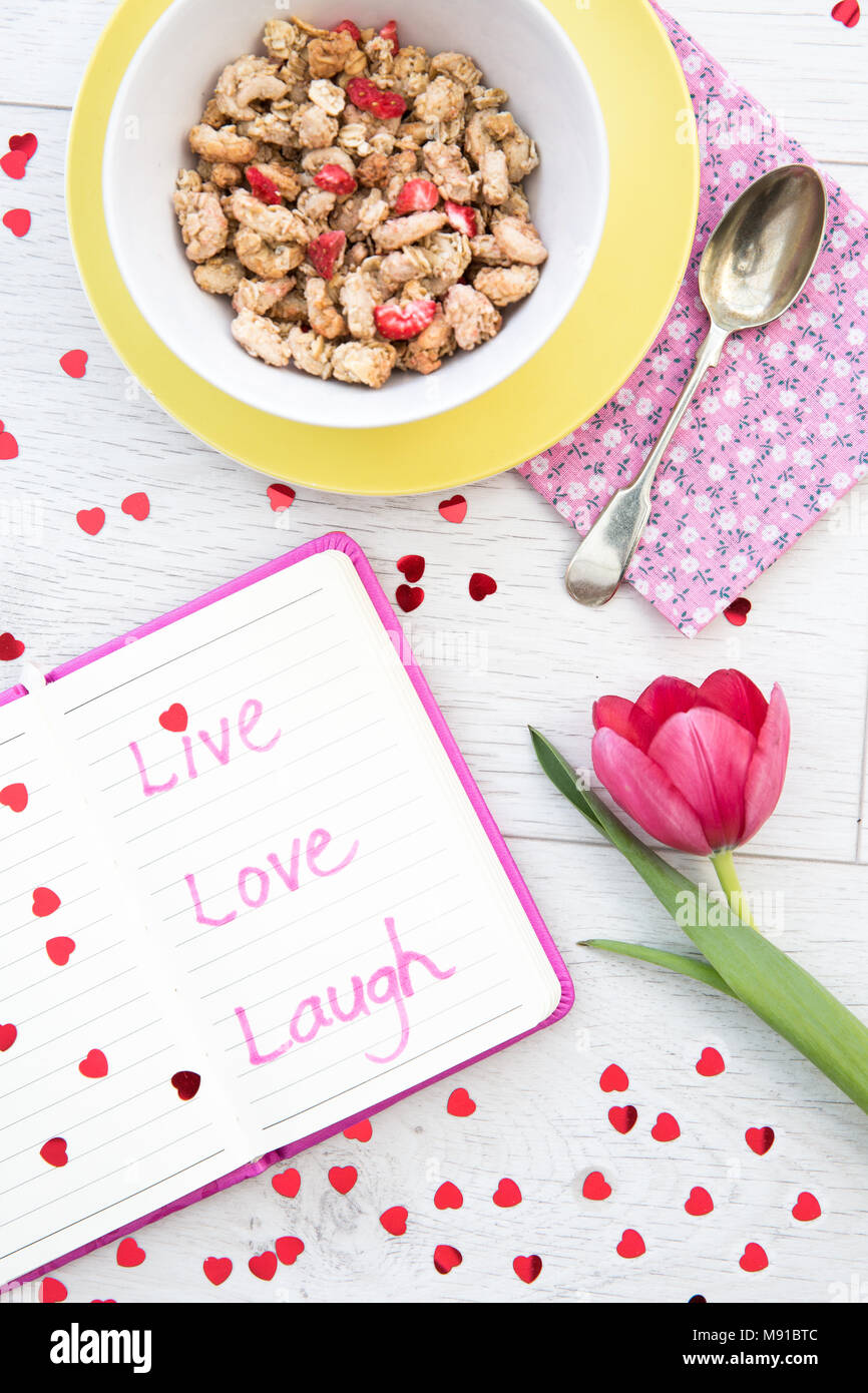 Looking down from above onto a white, wooden table top with a feminine breakfast of muesli, coffee and a notebook filled with inspiring quotes. Stock Photo