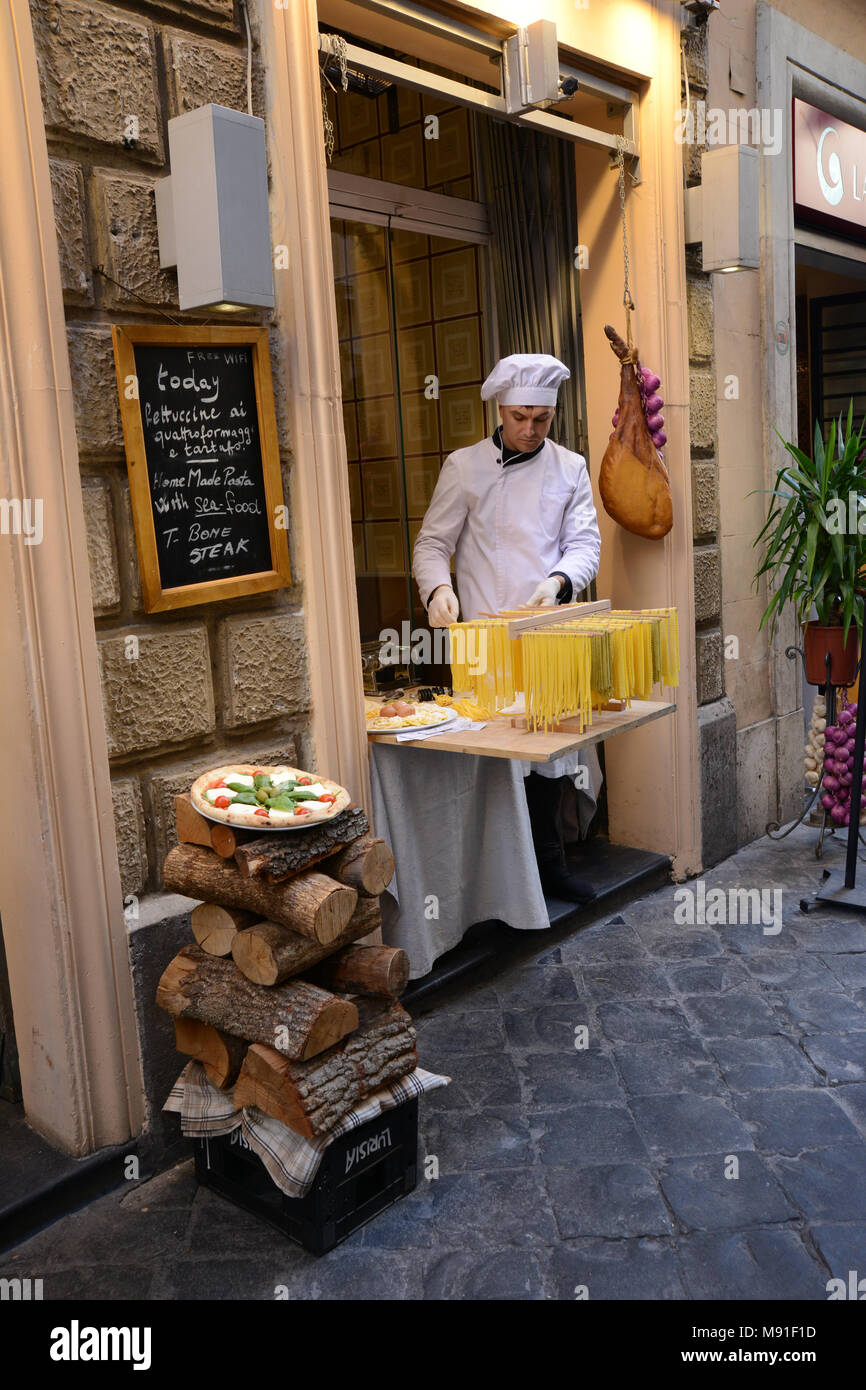 A chef in white overall and hat making pasta on the street in Rome with a pizza and a Palma Ham in the background. Stock Photo