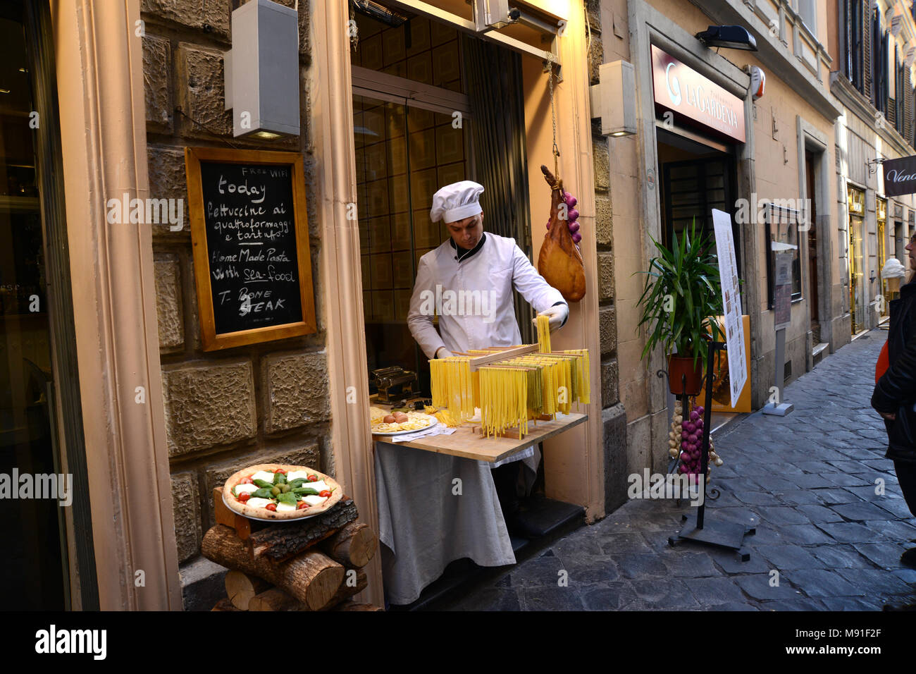 A chef in white overall and hat making pasta on Via della Croce in Rome with a pizza and a Palma Ham in the background. Stock Photo