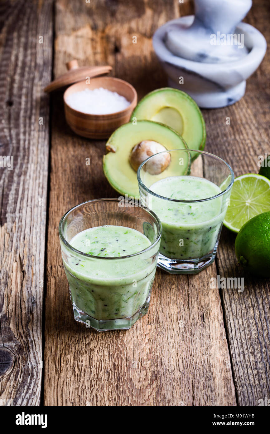 Guacamole dip style green vegetables smoothie in glasses with ingredients avocado and lime on rustic wooden table, savory healthy dairy drink with org Stock Photo