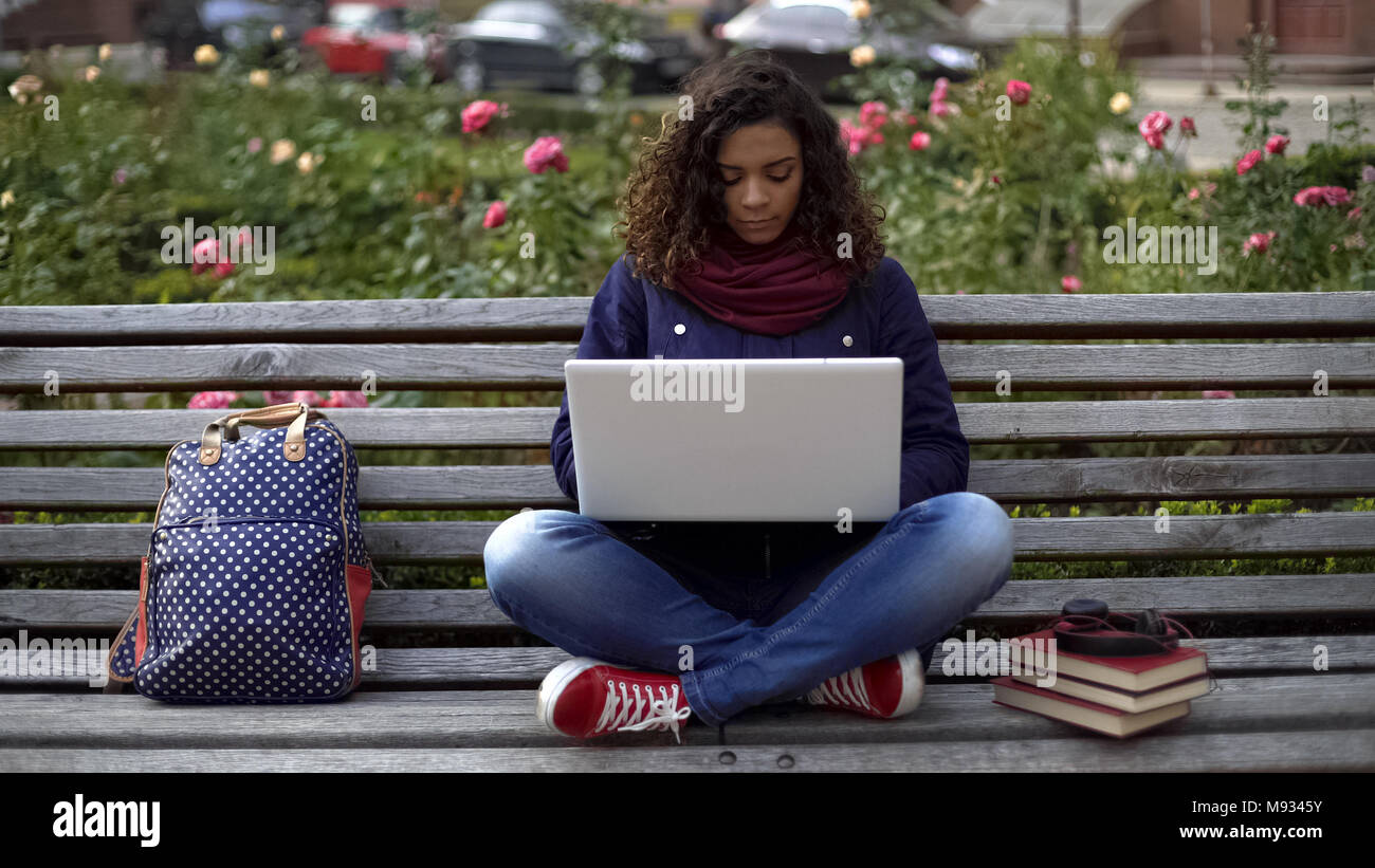 Young female student sitting on bench with laptop outdoors concentrated on study Stock Photo