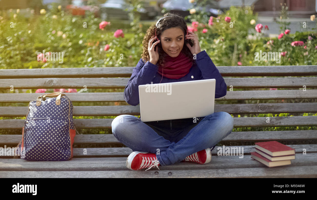 Smiling student in headphones sitting on bench, listening to favourite playlist Stock Photo