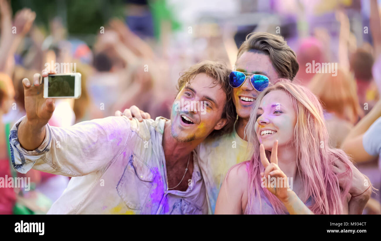 Excited friends covered in colorful powder at paint festival, posing for selfie Stock Photo