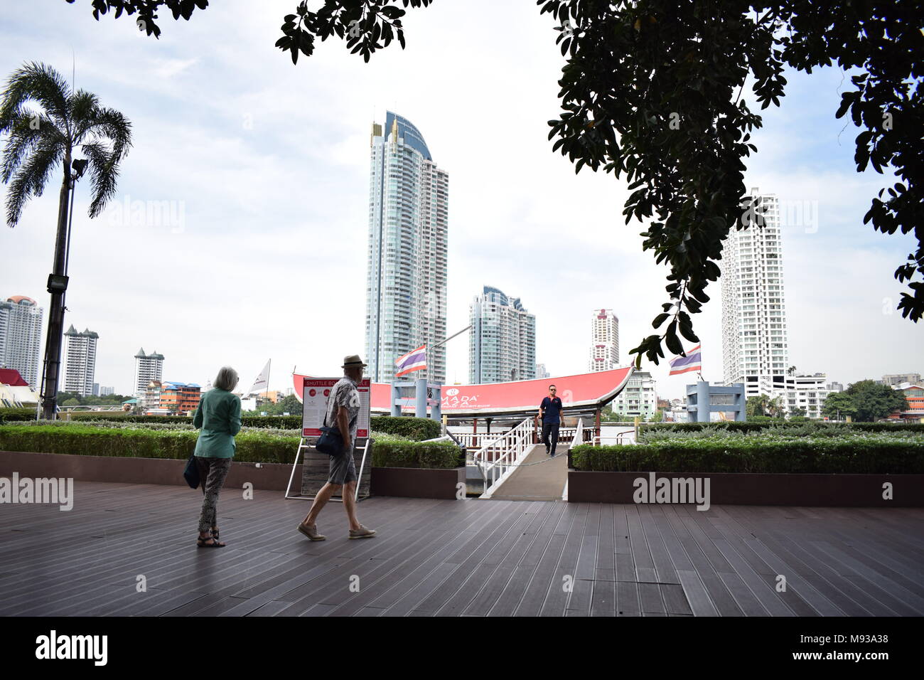 Beautiful picture of 2 tourists walking on a wooden deck before boarding on a river boat with skyscrapers in the background and trees in front. Stock Photo