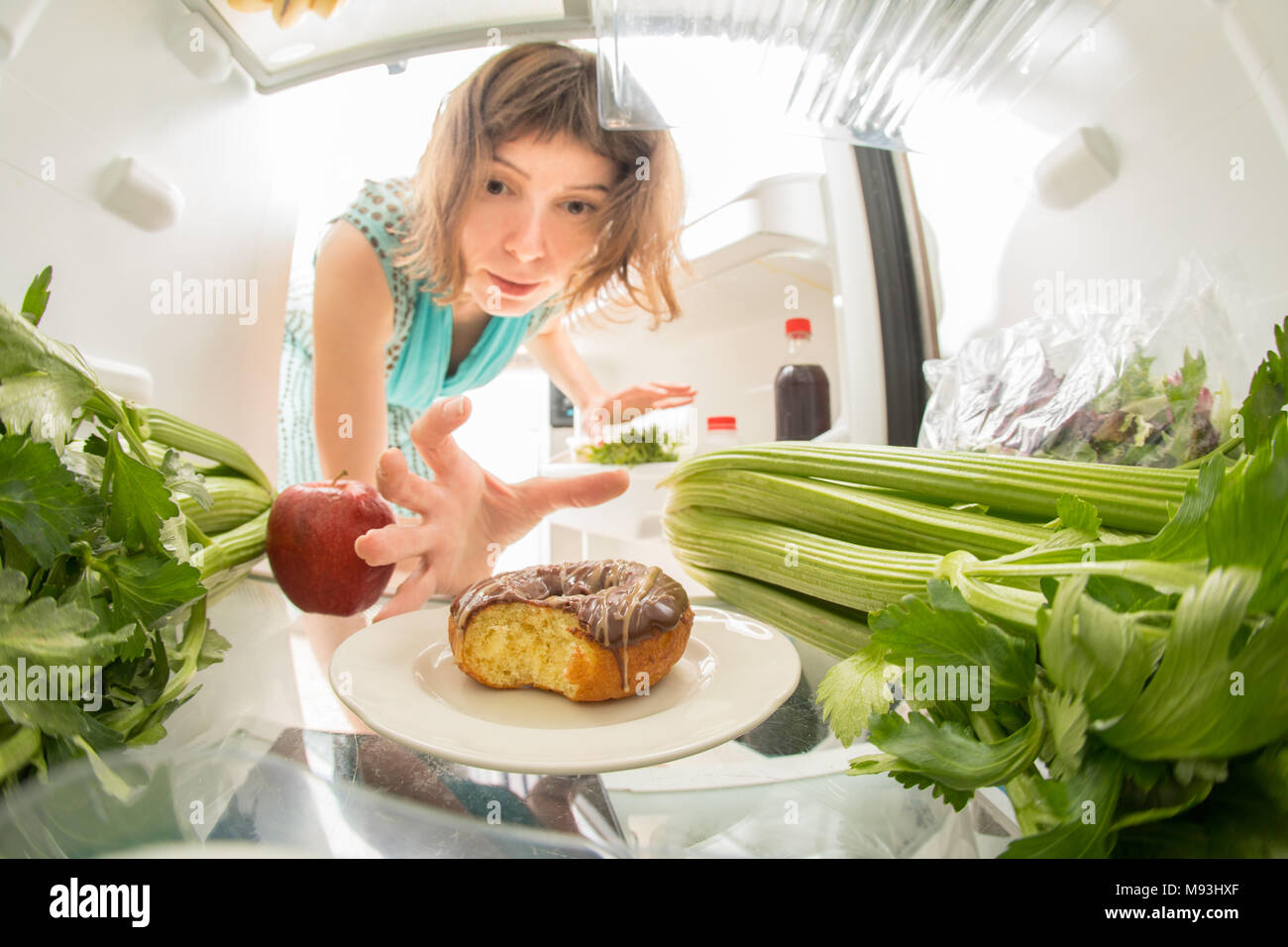 Diet struggle: A hand grabbing a donut from the open refrigerator full of greens. Stock Photo