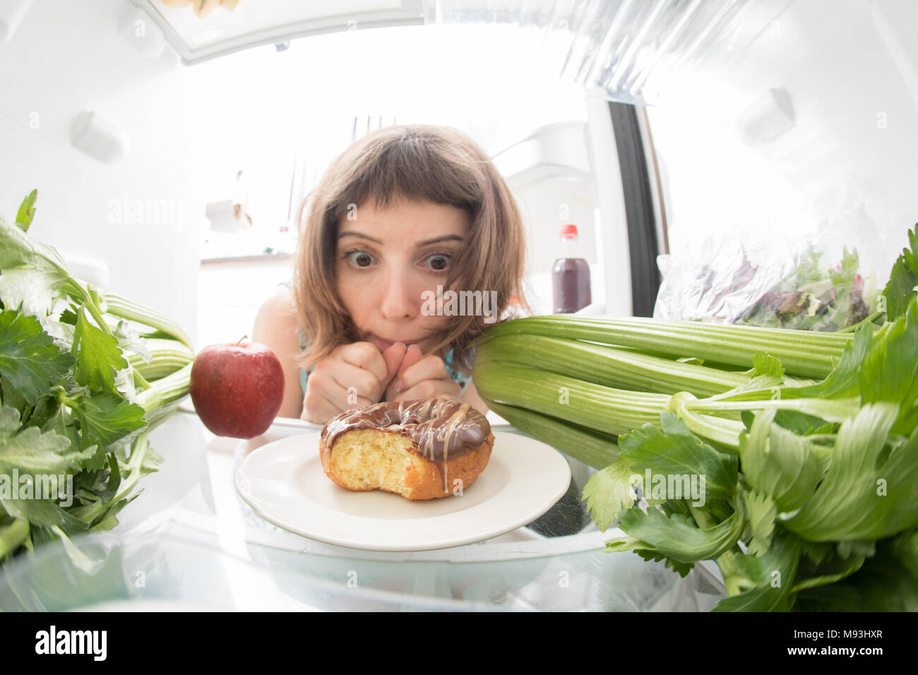 Diet struggle: A girl sadly looking at a donut inside a fridge full of healthy stuff. Stock Photo