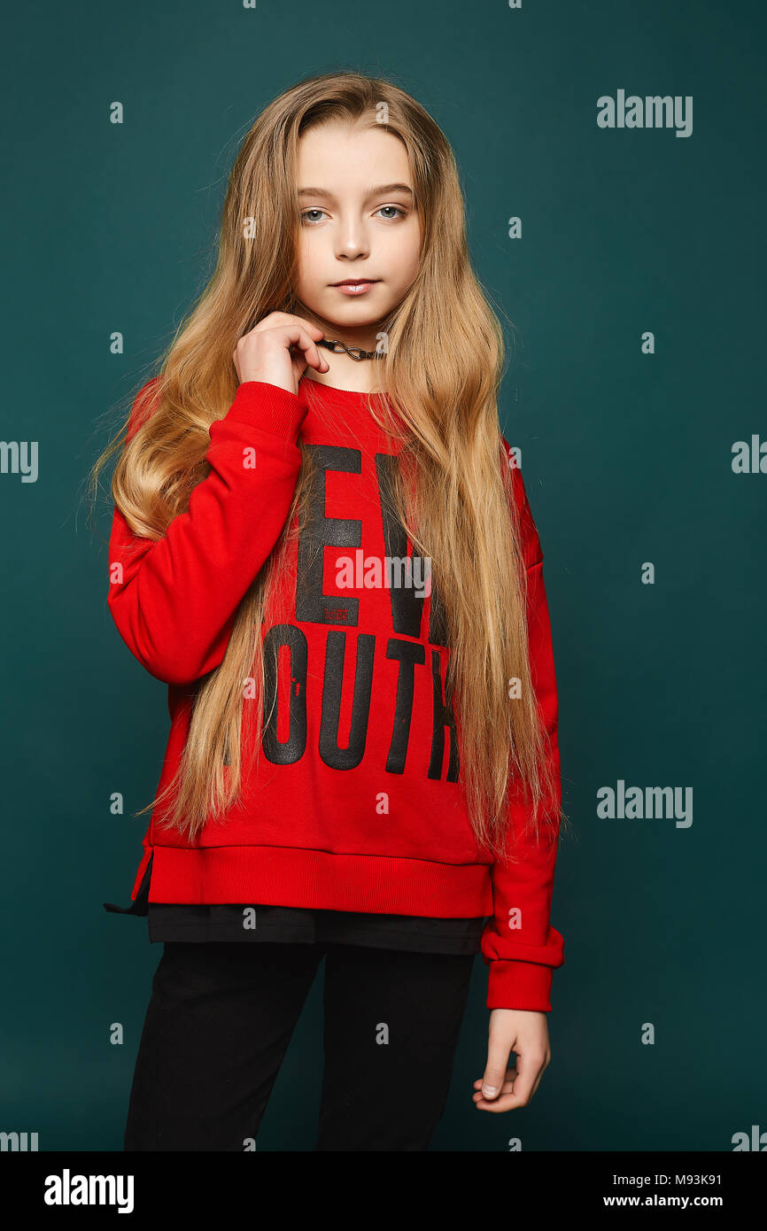 Stylish teen, beautiful young model girl with long blonde hair, posing at studio in jeans and red sweatshirt. Stock Photo