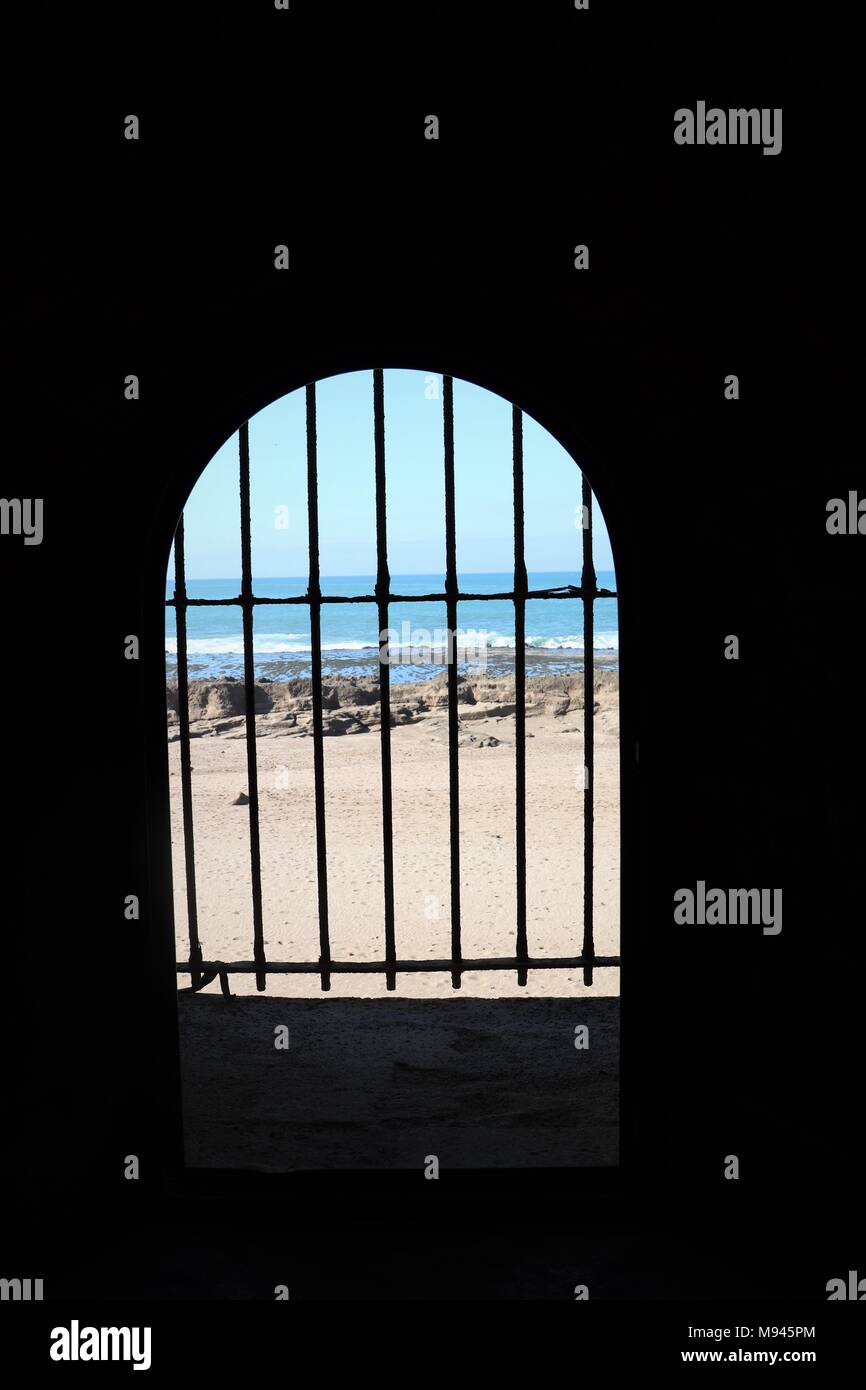 A sandy beach, rocks, rock pools and the sea are glimpsed through prison bars Borj Adoumoue Sale Morocco Stock Photo