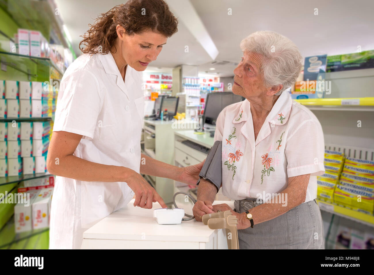 Young smiling female pharmaceutist taking old woman disabled,with crutches, blood pressure using sphygmomanometer in a real pharmacy backgroud Stock Photo