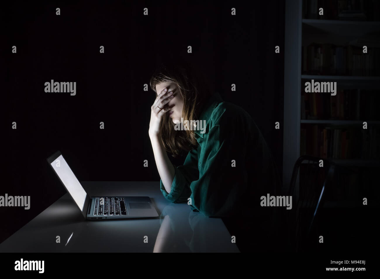 Tired and exhausted young woman hides eyes with hand at laptop pc late in the evening. Portrait of depressed female student or worker sitting in front Stock Photo