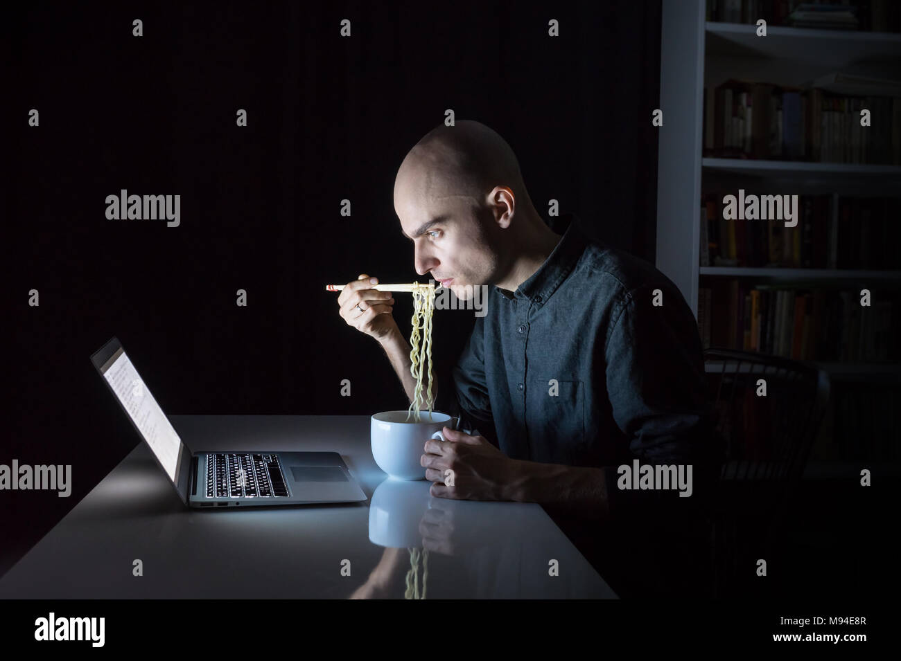 Young male at laptop computer eats instant ramen noodles with chopsticks late in the evening. Man working or studying online overtime at night has fas Stock Photo