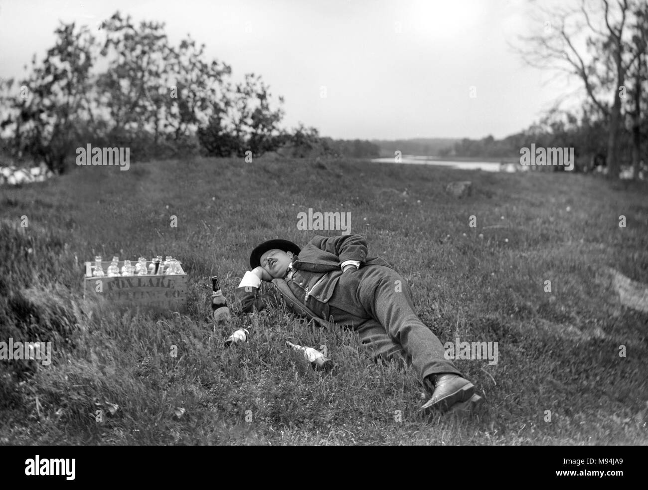 A man laying in the grass sleeps off an encounter with a case of beer from Fox Lake Brewery in Wisconsin, ca. 1905. Stock Photo