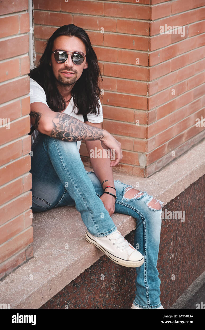 Attractive young man wearing sunglasses and long hair relaxing in urban background.  Sunglasses  male model with long hair and tattoos on hand. Stock Photo
