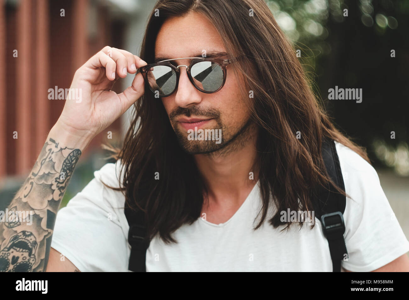 Sunglasses man model with long hair, beard and tattoos. Stylish male  portrait wearing white t-shirt and backpack, fixing eyewear with hand on  urban ba Stock Photo - Alamy