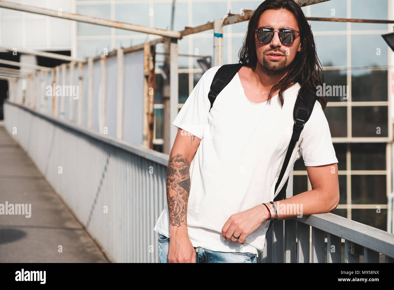 Young attractive bearded man with tattoos and long hair wearing sunglasses  white shirt and backpack posing on urban background Stock Photo  Alamy