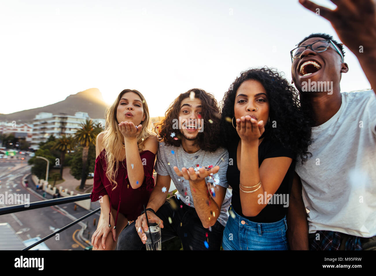 Group of friends blowing confetti at rooftop party. Men and women having a great time together on terrace party. Stock Photo
