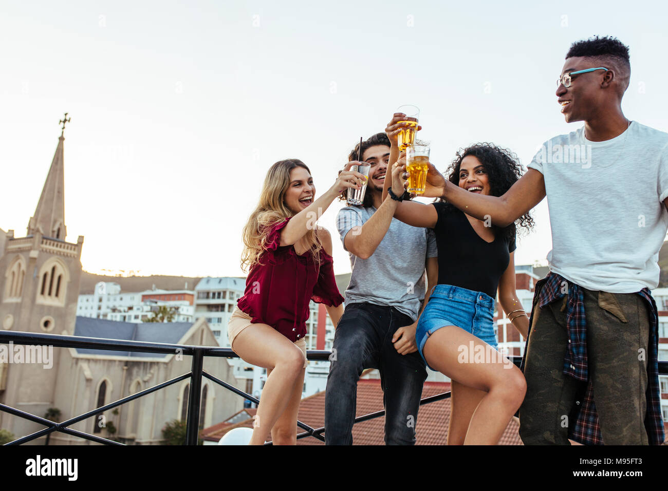 Friends and drinks. Young cheerful people cheering with beer and smiling on rooftop party. Men and women toasting drinks on terrace. Stock Photo