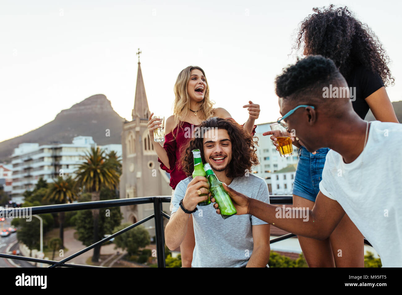Happy young men toasting beers with girls laughing and enjoying at the back on terrace. Group of friends having rooftop party with drinks. Stock Photo