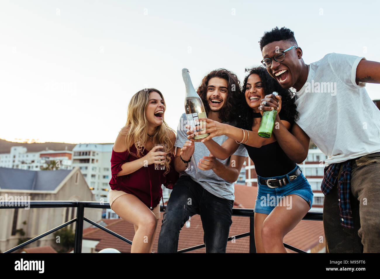 Outdoor shot of young woman opening a bottle of champagne with friends. Young people having champagne at rooftop party. Stock Photo