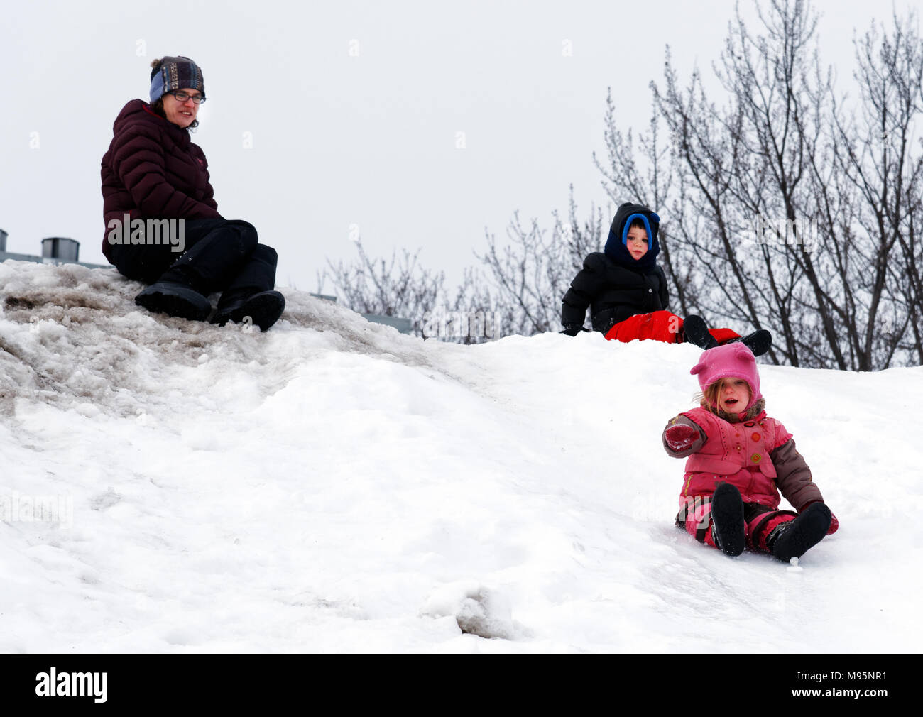 A little girl sliding on her bum in the snow, with her mum and brother looking on Stock Photo