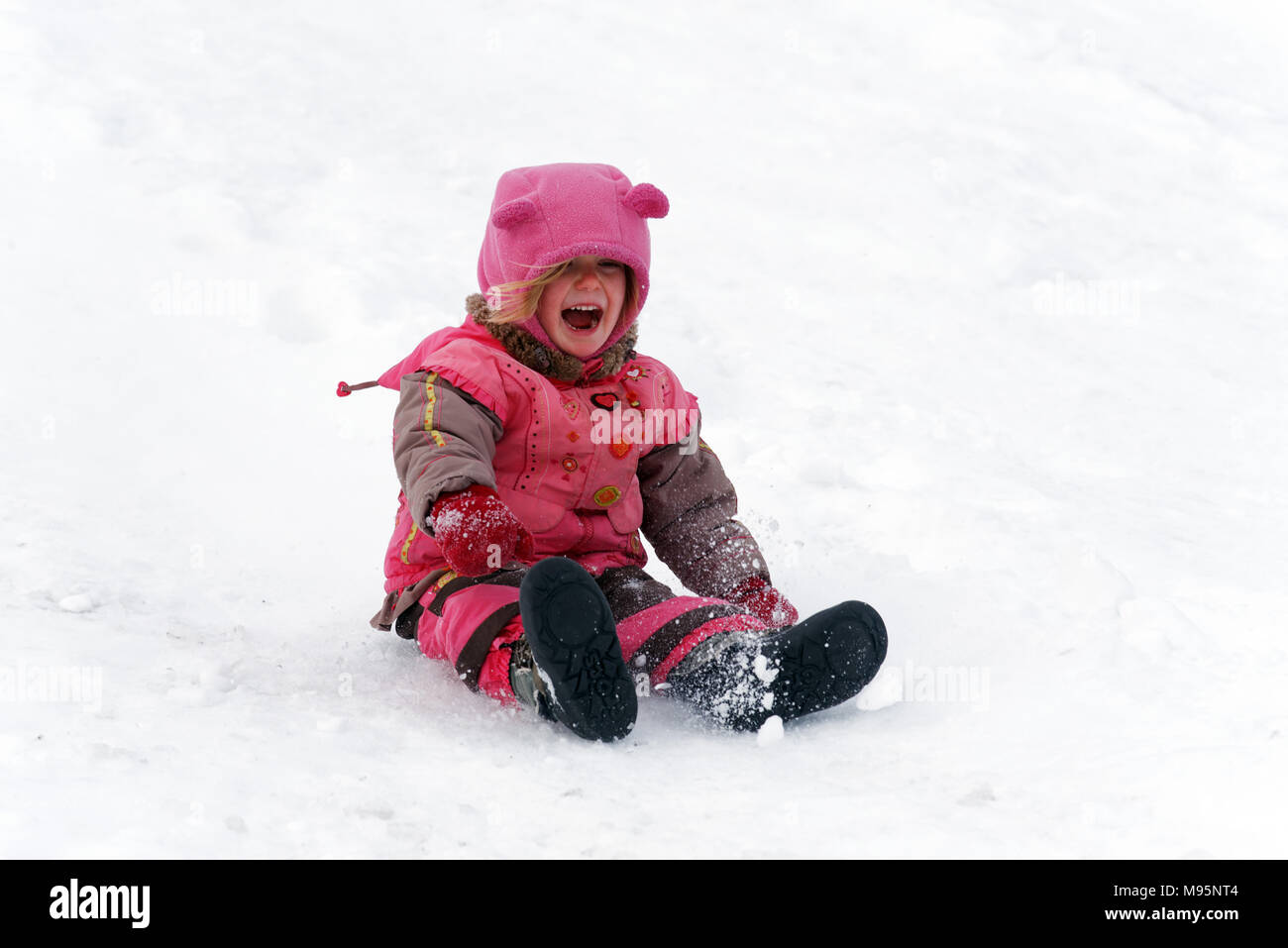 A laughing three year old girl sliding on her bum in the snow in Quebec Canada Stock Photo