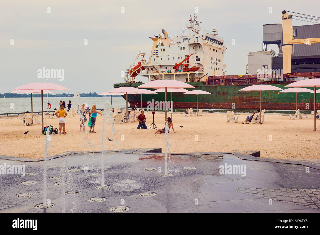 Pink umbrellas and holidaymakers on urban beach next to Lake Ontario with bulk cargo carrier Mandarin in the background, Toronto, Ontario, Canada Stock Photo