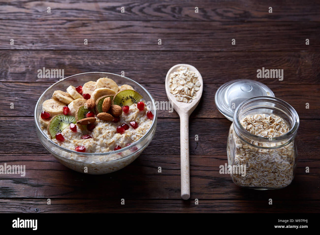 Composition with transparent bowl of oatmeal porrige with banana, kiwi, almond, pomergranate, dry oatmeal in glassware and wooden spoon on vintage woo Stock Photo