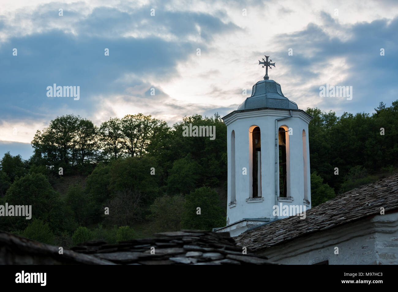 Lit dome of orthodox church with decorated cross and brass bell at evening, village of Dolen, Rhodope mountain, Bulgaria with cloudy sky background Stock Photo