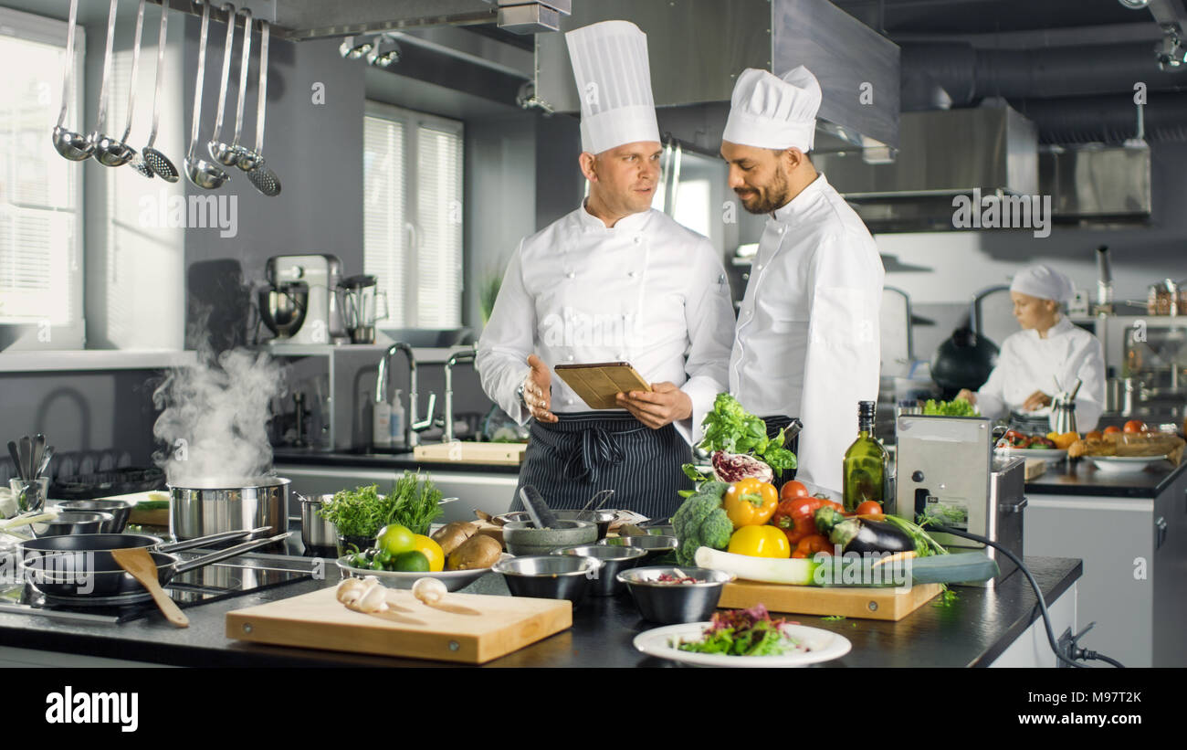 Two Famous Chefs Exchange Ideas About Video Blog Recipe they Watching on Tablet. They are Working in Big Restaurant Kitchen. Stock Photo