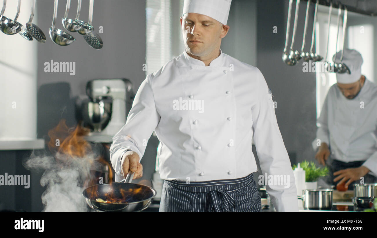 Professional Chef Fires up Oil on a Pan. Flambe Style Cooking. He Works in a Modern Kitchen with Lots of Ingredients are Around. Stock Photo